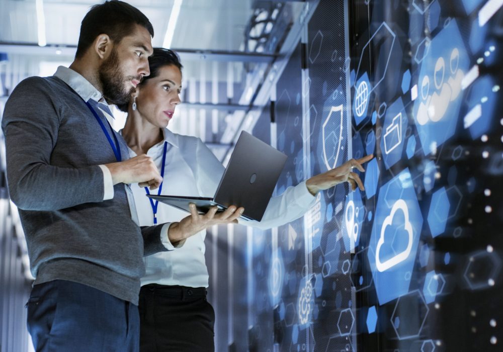 Male IT Specialist Holds Laptop and Discusses Work with Female Server Technician. They're Standing in Data Center, Rack Server Cabinet with Cloud Server Icon and Visualization.