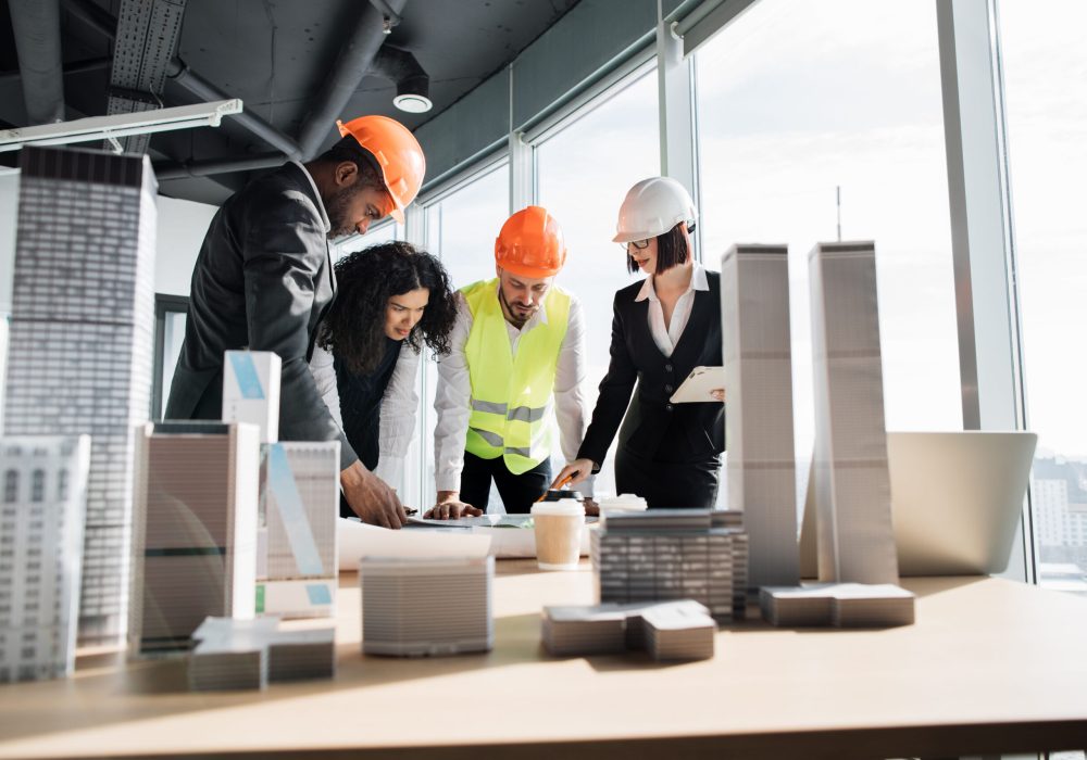 Group of four multiracial people architects standing near desk with lots of blueprints and architecture design of buildings with residential project maquette at office with panoramic windows.